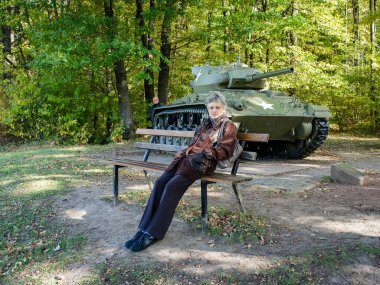 Neşeli olgun kadın fotoğrafçıya gülümsedi. Tankın yanındaki sonbahar şehir parkında dinleniyor..
