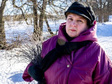 Kasketli ve ceketli olgun bir kadın kış günü nehir kenarındaki bir parkta yürüyor.