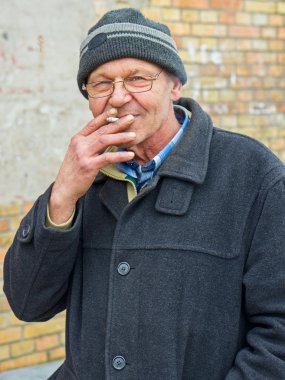 Yaşlı gri saçlı adam gözlük ve açık havada duman exhaling ve dikkatle kameraya bakarak kış woodland sigarada zevk örme şapkası giyiyor