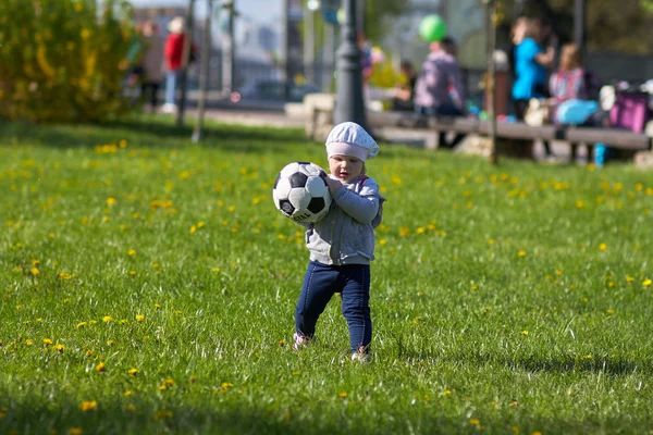 Liten flicka baby blond utomhus. Aktiva fotbollsfan körs på fältet grönt gräs med boll — Stockfoto