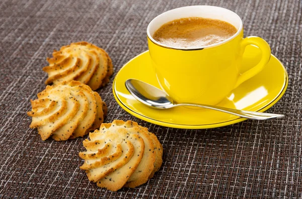 Three shortbread cookies with poppy seeds, coffee espresso in yellow cup, spoon on saucer on dark mat