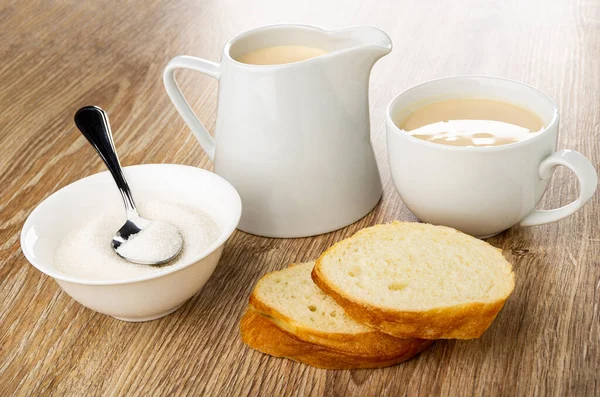 Teaspoon in white bowl with sugar, pitcher and cup with fermented baked milk, slices of bread on wooden table