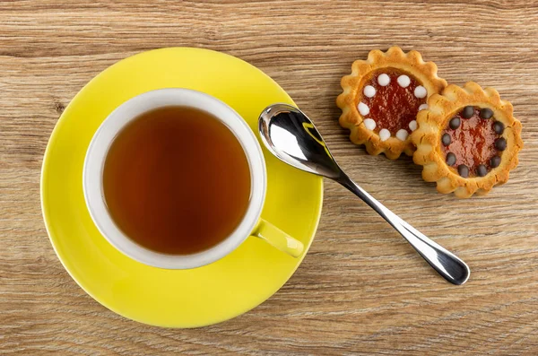 Cup of tea on yellow saucer, cookies with filling from orange jam and chocolate, teaspoon on wooden table. Top view