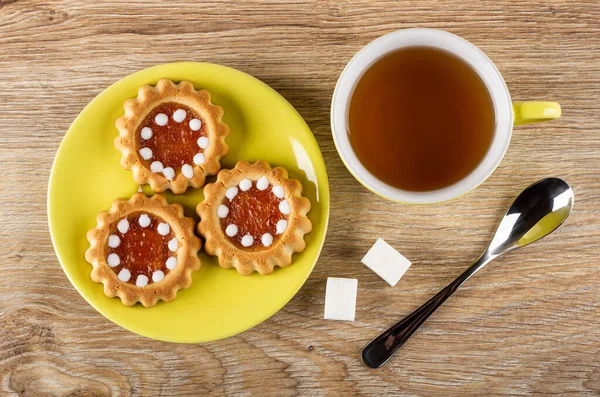 Cookies with filling from orange jam and chocolate in yellow saucer, sugar cubes, teaspoon, cup of tea on wooden table. Top view