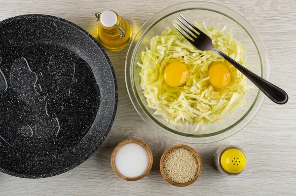 Frying pan with vegetable oil, bottle of vegetable oil, eggs in chopped cabbage, fork in transparent bowl, bamboo bowls with sesame and salt, pepper shaker on wooden table. Top view