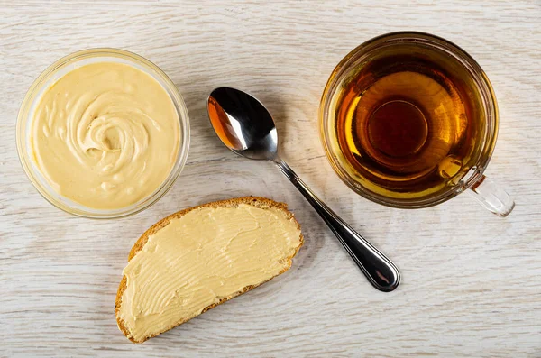 Bowl with dairy peanut paste, teaspoon, sandwich with peanut butter, cup of tea on wooden table. Top view