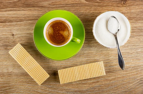 Wafers, coffee espresso in cup on green saucer, spoon in white bowl with sugar on brown wooden table. Top view