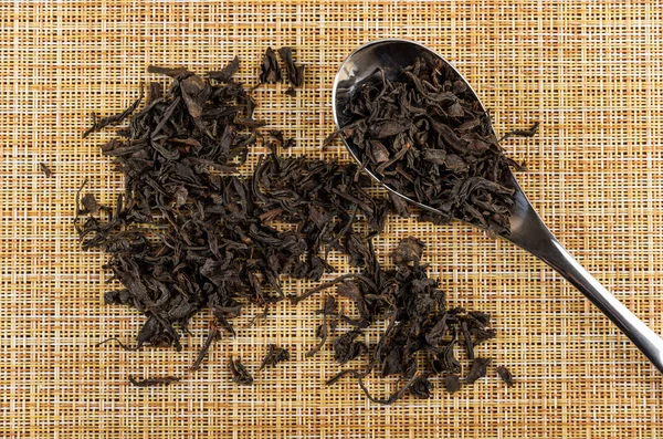 Scattered leaf black tea, leaf tea in metallic spoon on brown mat. Top view