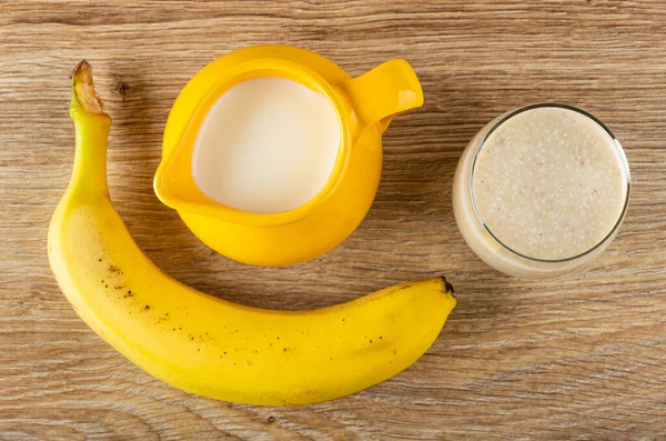 Banana, yellow pitcher with milk, glass of dairy cocktail on wooden table. Top view