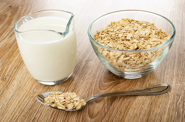 Pitcher with milk, transparent bowl with raw oat flakes, metallic spoon with oat flakes on wooden table