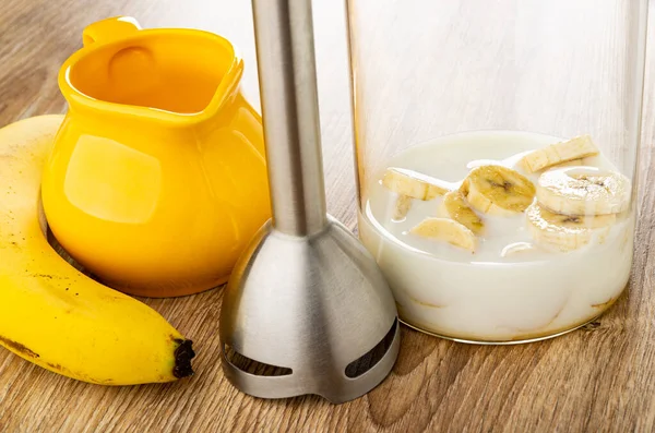 Banana, yellow pitcher with milk, electric blender, slices of peeled banana with milk in transparent glass on wooden table