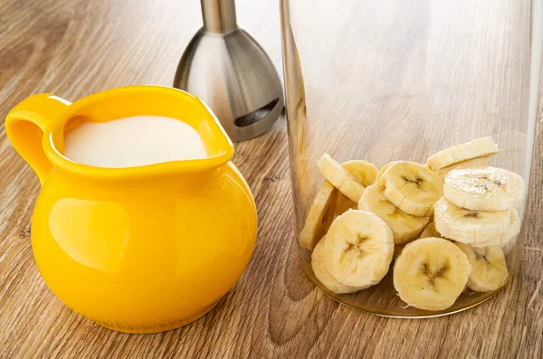 Electric blender, yellow pitcher with milk, slices of peeled banana in transparent glass on wooden table