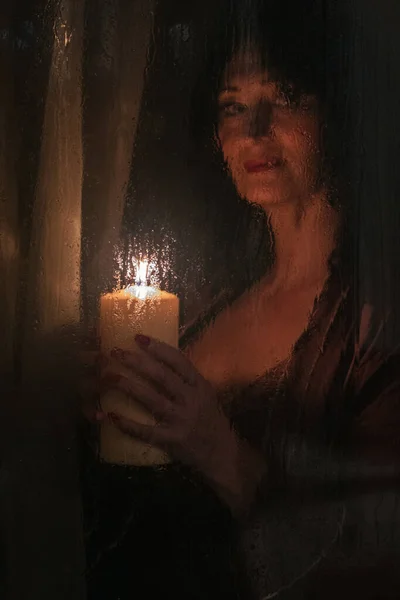 美丽的女人在湿透的玻璃杯后面拿着一支大蜡烛 点着雨滴 — 图库照片