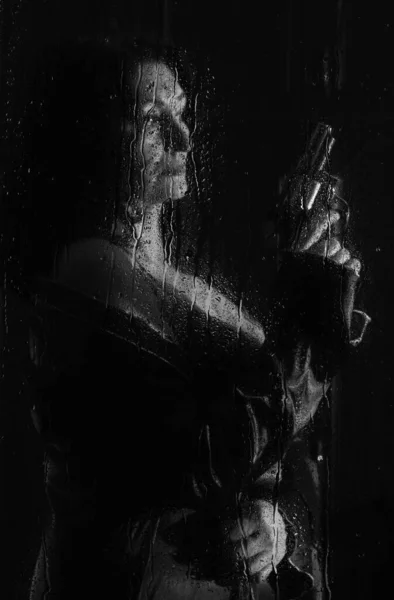 beautiful woman with a revolver in her hands behind a wet glass with raindrop