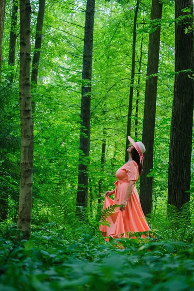 長いサンゴのドレスを着た美しい大人の女性と夏の森を歩く広い帽子 — ストック写真