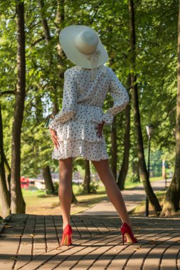 slender woman in a short summer dress, wide hat and high-heeled shoes in the morning in the park