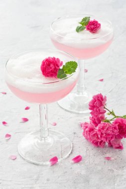 İki şarap kadehinde gül çiçekleriyle pembe kokteyl protein köpüklü gül ve gri beton arka planda nane yaprağı.