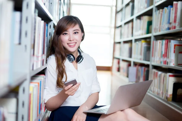 Estudiante femenina usando portátil y teléfono móvil en la biblioteca — Foto de Stock
