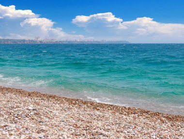 Güneşli bulutlu bir gökyüzünün altında çakıl taşı plajı ve deniz dalgaları