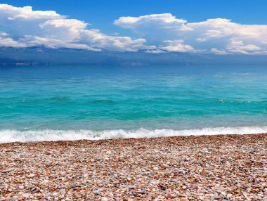 Güneşli bulutlu bir gökyüzünün altında çakıl taşı plajı ve deniz dalgaları