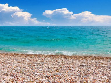 Güneşli bulutlu bir gökyüzünün altında çakıl taşı plajı ve deniz dalgaları