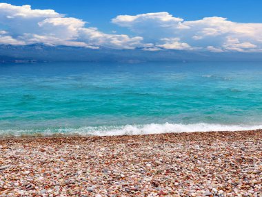 Güneşli bulutlu bir gökyüzünün altında çakıl taşı plajı ve deniz dalgaları