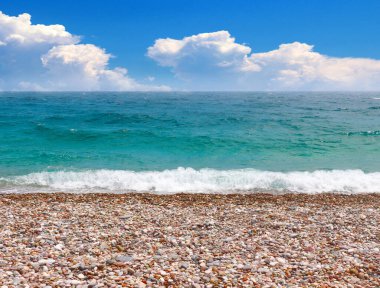Güneşli bulutlu bir gökyüzünün altında çakıl taşı plajı ve deniz dalgaları