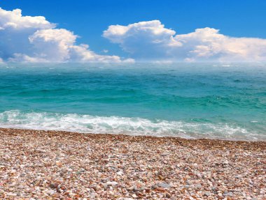 Güneşli bulutlu bir gökyüzünün altında çakıl taşı plajı ve deniz dalgaları