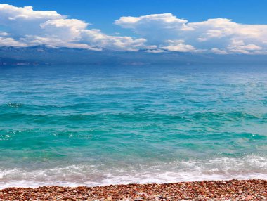 Güneşli bulutlu bir gökyüzünün altında çakıl taşı plajı ve deniz dalgaları