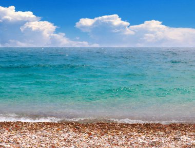 Güneşli bulutlu bir gökyüzünün altında çakıl taşı plajı ve deniz dalgaları