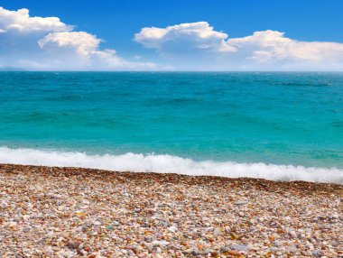 Güneşli bulutlu bir gökyüzünün altında çakıl taşı plajı ve deniz dalgaları