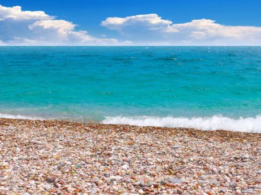 Güneşli bulutlu bir gökyüzünün altında çakıl taşı plajı ve deniz dalgaları