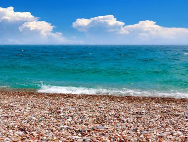 Güneşli bulutlu bir gökyüzünün altında çakıl taşı plajı ve deniz dalgaları