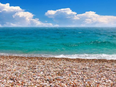 Güneşli bulutlu bir gökyüzünün altında çakıl taşı plajı ve deniz dalgaları
