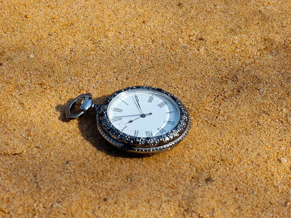 an antique pocket watch in a precious metal case lies on the sand as a symbol of time