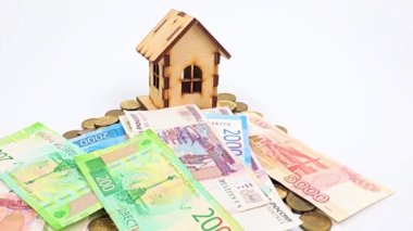Ahşap bir ev, metal ruble yığını üzerinde duruyor ve kağıt gagalar düşüyor.
