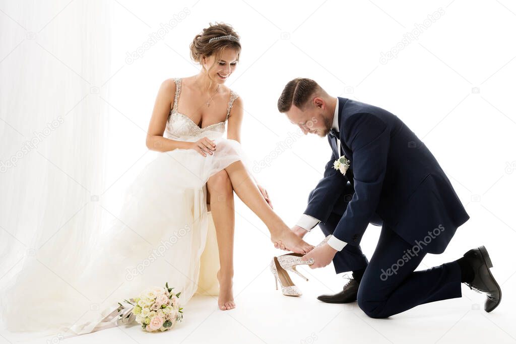 Fashion Wedding Couple. Groom putting on Cinderella Shoes on Bride Leg. Elegant Bridal Preparation. Isolated white