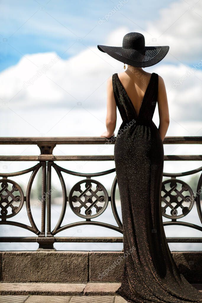 Woman Back Side View in Hat and Evening Dress Outdoor. Fashion Model Rear View looking away. Luxury Lady in long Gown at Promenade looking at Sky