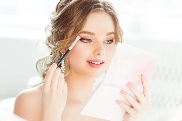 Bride Wedding Makeup. Bridal Make up. Beautiful Woman looking at Hand Mirror and putting Eye Shadow Eyebrow Self Make up in White Room