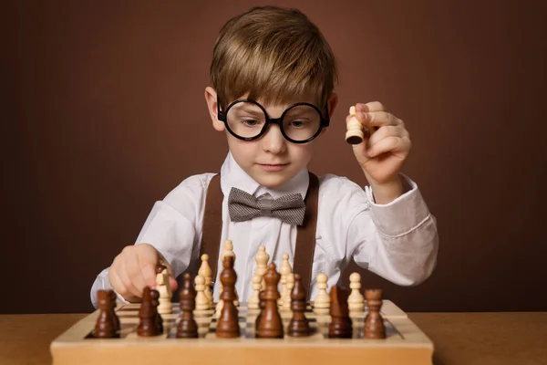 チェスゲームをしている小さな子供 チェスボードの横にあるメガネのインテリジェントな小さな男の子 子供の教育と開発 ブラウンスタジオの背景 — ストック写真