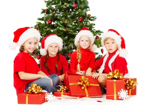 Niños de Navidad en el sombrero de Santa con regalos sentados bajo el árbol de Navidad, fondo blanco —  Fotos de Stock