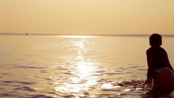 Menino brincando com uma bola no mar em um pôr do sol — Vídeo de Stock
