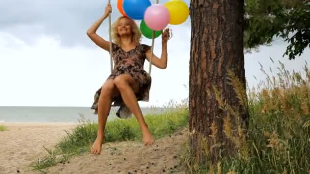 Dziewczyna z kolorowych balonów jest zabawy na huśtawce pod drzewem na plaży. — Wideo stockowe