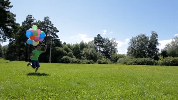 Glücklicher Junge rennt mit vielen bunten Luftballons auf grünem Gras — Stockvideo