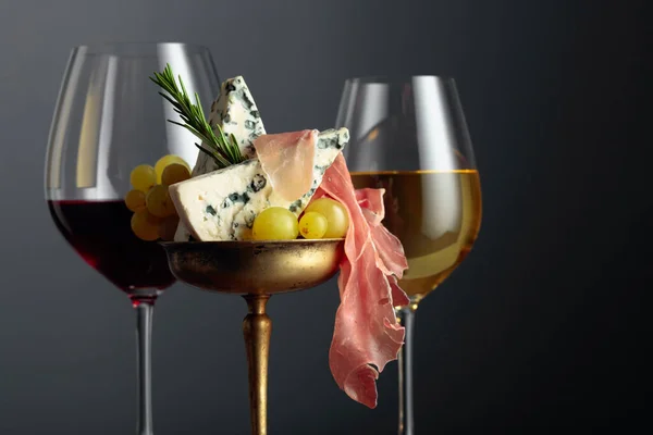 Red and white wine, blue cheese, prosciutto, grapes, and rosemary on a black background. Simple and tasty food. Copy space.