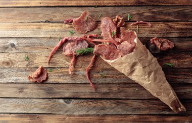 Eski ahşap bir masada, kağıt bir torbada biberiyeli baharatlı kurutulmuş et dilimleri. Üst Manzara. Boşluğu kopyala.