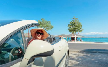 Şapka ve güneş gözlüğü takan orta yaşlı çekici bir kadın. Güzel bir deniz manzarasının arka planında üstü açık bir arabada gülümseyen bir kadın..