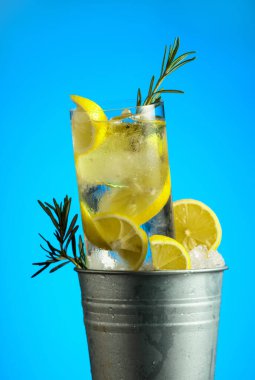 Buzlu, limonlu ve biberiyeli cin tonik kokteyl. Mavi arka planda buz olan metal bir kovada..