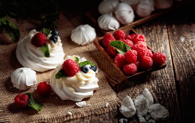 Pavlova 'ya ahududu, yaban mersini ve nane şekerini eski ahşap bir masaya koyun. Pavlova tatlısı ve kırsal bir arka planda küçük kremalar.