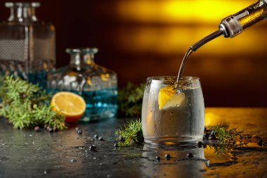 Kokteyl cini ve tonik, ardıç dalları ve siyah bir masada limon dilimleri. Cin şişeden buz ve limon dilimli bardağa dökülür. Sarı arka ışık.